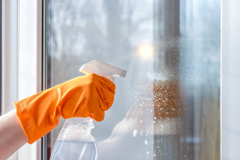 Mano con guanto arancione pulisce finestra con spray.